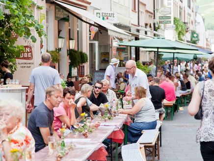 Lange Tafel Das Bild zeigt ein Ausschnitt von der Langen Tafel in Zell an der Mosel. Man erkennt viele Personen, die an Tischen vor Restaurants und Cafés sitzen und sich unterhalten.