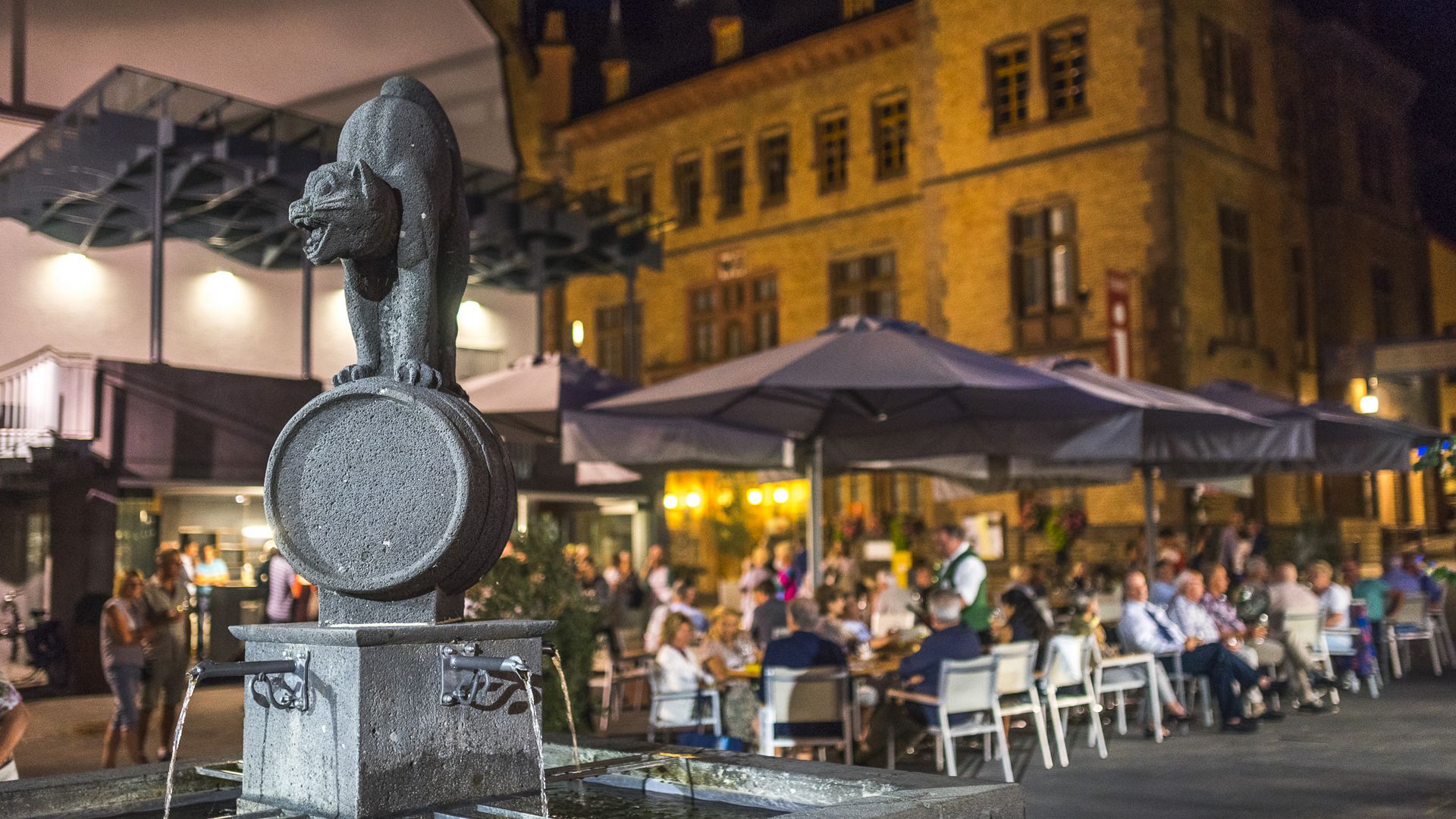 Zeller Schwarze Katz Brunnen Das Bild zeigt den Zeller Schwarze Katz Brunnen beim Moselabend. Im Hintergrund erkennt man den Marktplatz, auf dem viele Tische mit Schirmen aufgebaut sind, an denen mehrere Personen sitzen.