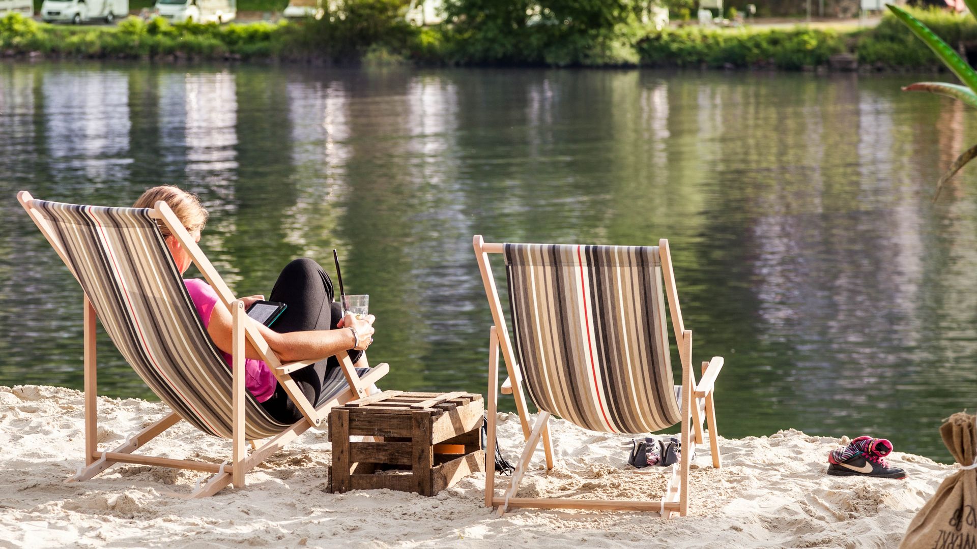 Sommertipps Das Bild zeigt eine Frau, die in einem Liegestuhl am Moselufer sitzt, an dem Sand verteilt ist. Dabei liest sie und hält ein Getränk in ihrer Hand.