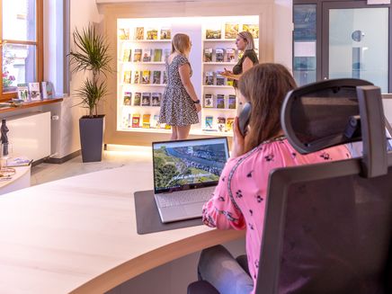 Tourist Information Auf dem Bild befindet sich eine Dame hinter dem Counter die telefoniert. Und vor der Prospektwand schauen sich zwei Frauen die Prospekte an.