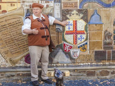Riwigmännchen Auf dem Bild befindet sich ein Herr in einem Kostüm. Er ist der Zeller Stadtgeist - das Riwigmännchen. Er steht vor dem Zeller Stadtwappen und erzählt einiges dazu.