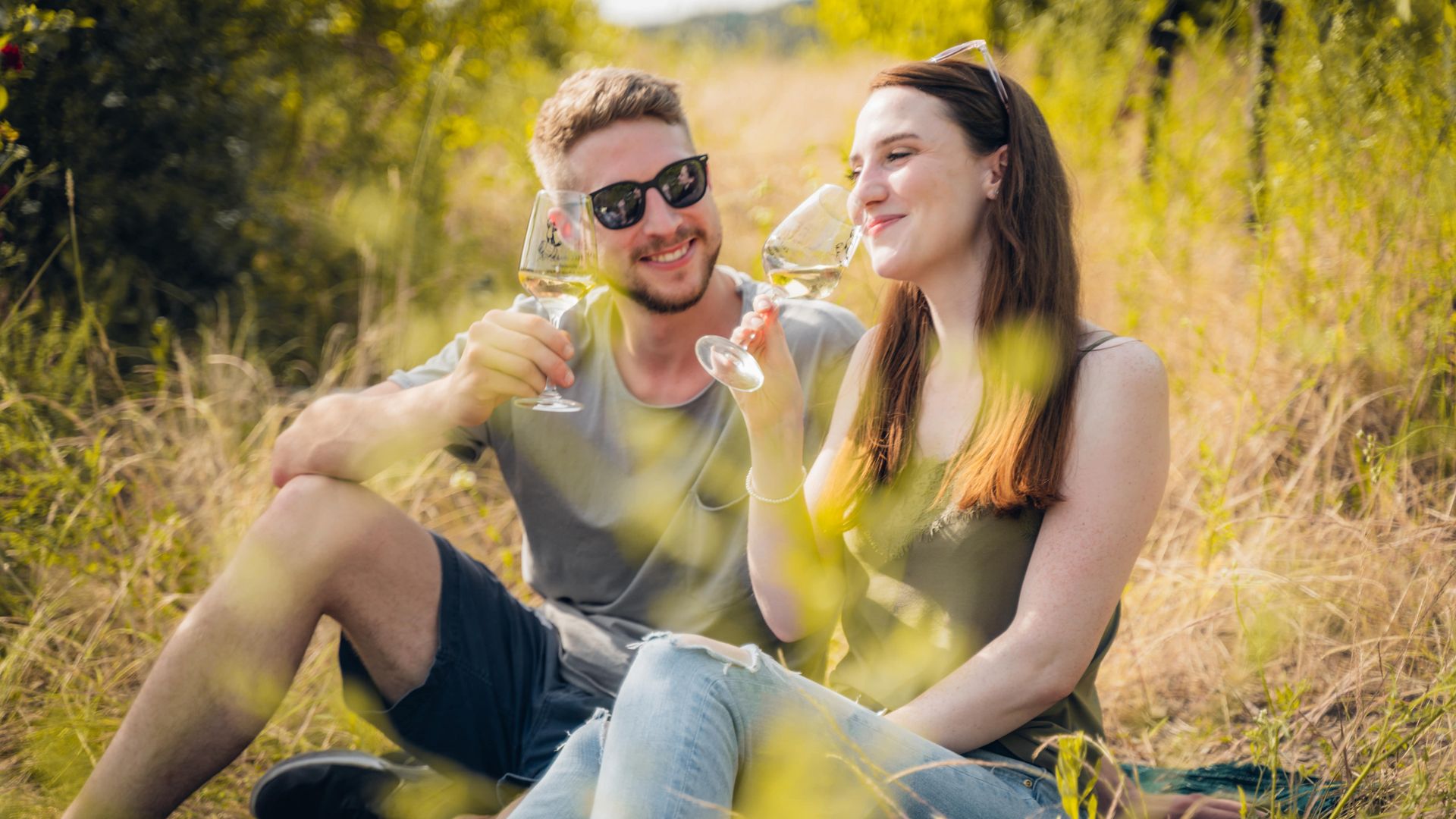 Wein erleben Auf dem Bild befinden sich zwei junge Leute, die auf einer Picknickdecke sitzen und ein Glas Wein trinken.