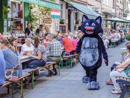 Lange Tafel Das Bild zeigt eine Aufnahme von der Langen Tafel in der Stadt Zell. Rechts und links sitzen Personen, die sich unterhalten sich und in der Mitte geht eine Person, verkleidet als die Zeller Schwarze Katz.