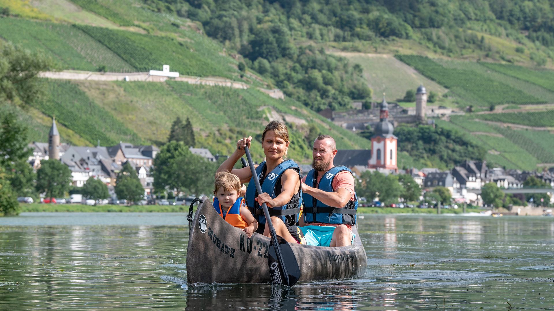 Das Bild zeigt eine Familie die auf der Mosel Kanu fährt. Im Hintergrund befindet sich die Altstadt von Zell an der Mosel und die Weinberge.