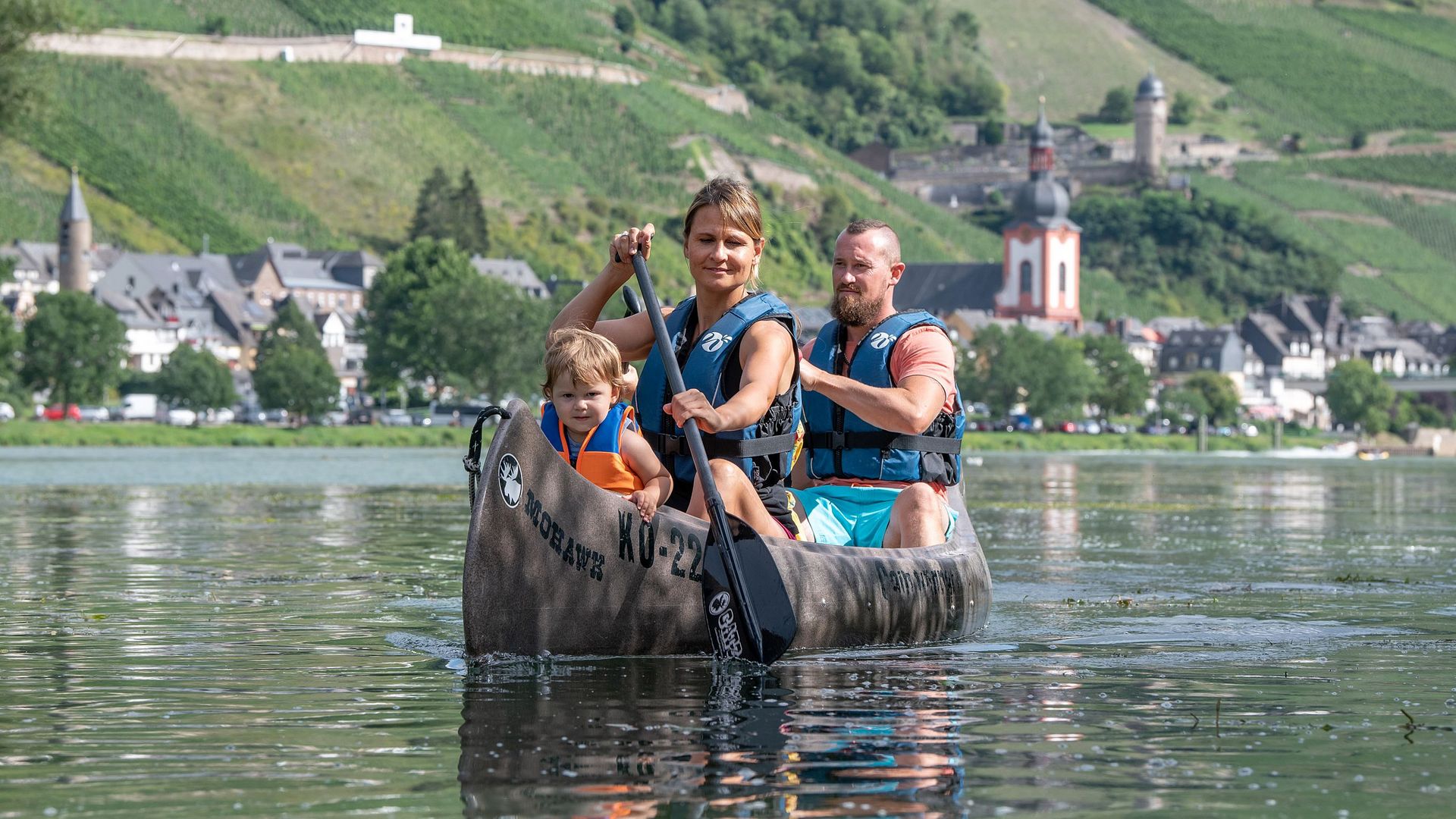 Kanu fahren Das Bild zeigt eine Familie die auf der Mosel Kanu fährt. Im Hintergrund befindet sich die Altstadt von Zell an der Mosel und die Weinberge.