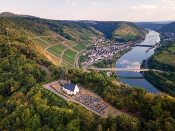 Das Bild ist eine Drohnenaufnahme. Auf dem Bild befindet sich die Kapelle und der Höhenfriedhof auf Anhöhe. Und unten ist der Fluss mit der Brücke und der Schleuse und der Ort Neef.