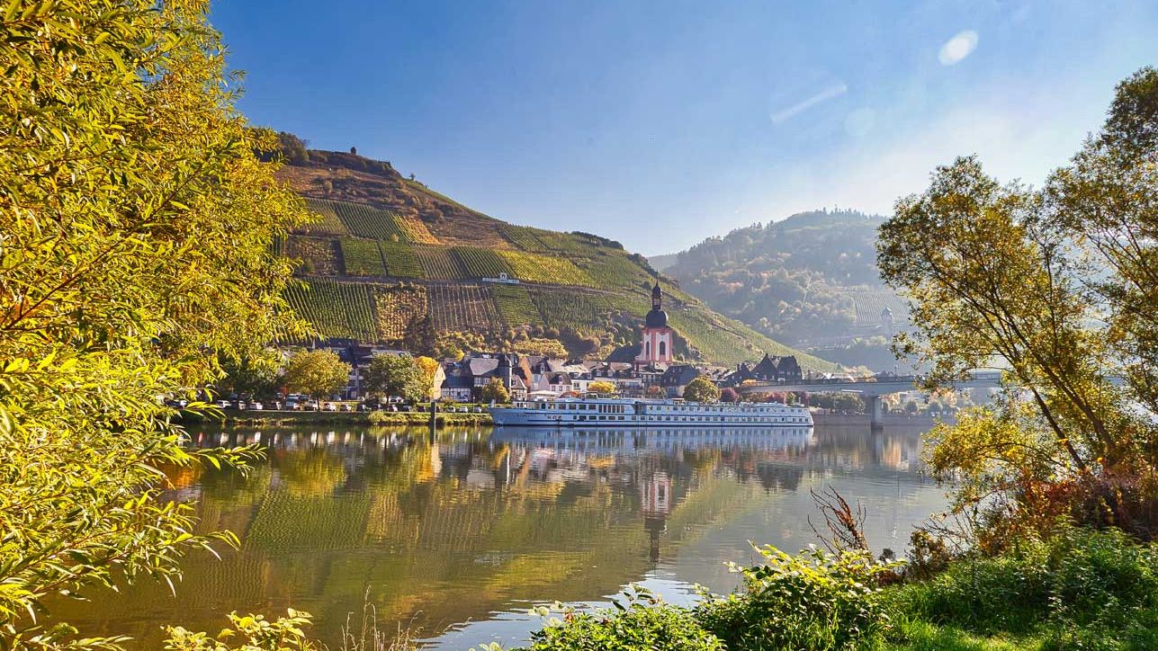 Schifffahrten Das Bild zeigt ein Kreuzfahrtschiff auf der Mosel bei schönem Wetter, welches vor der Stadt Zell steht. Im Hintergrund befinden sich die St. Peter Kirche, die Fußgängerbrücke sowie die Weinberge. Das Bild wurde von dem Moselufer in Zell-Kaimt aufgenommen, wodurch am Rand der Aufnahme Abschnitte von Bäumenzu erkennen sind.