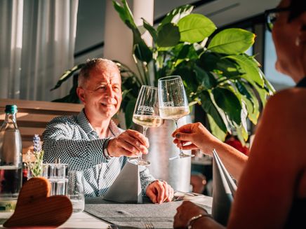 Zusammen anstoßen Auf dem Bild befinden sich zwei Personen die an einem Tisch im Restaurant sitzen und mit einem Glas Wein anstoßen.