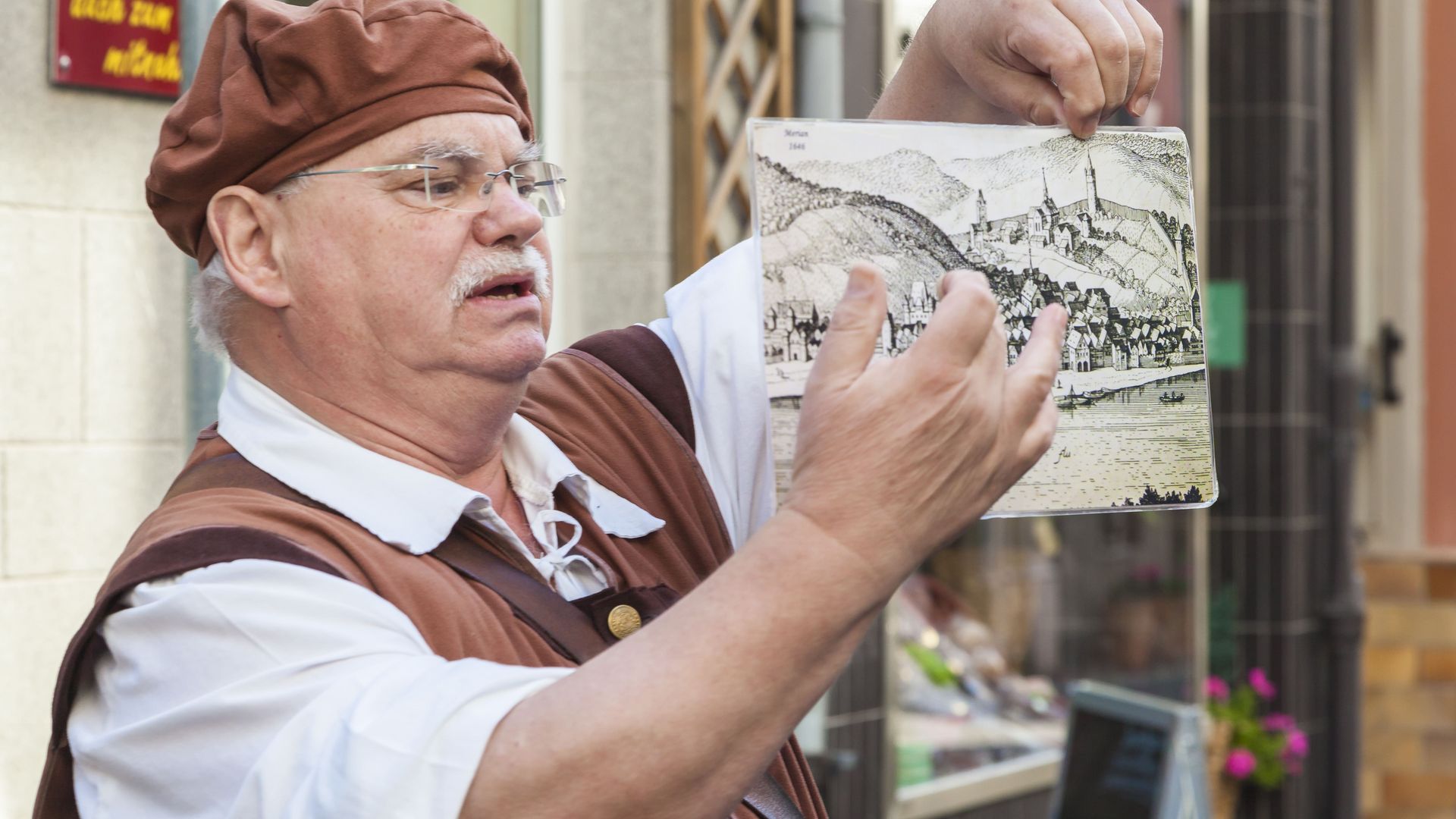 Riwigmännche in Zell (Mosel) In the picture there is an elderly gentleman in a costume. He points to a picture depicting the old town of Zell many years ago. He is explaining past stories to tourists as the town ghost of Zell.