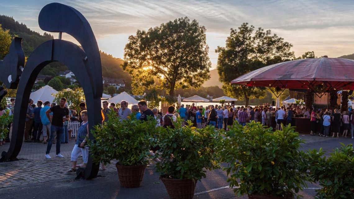 Weinfest  Das Bild zeigt eine Aufnahme von dem Weinfest in Zell an der Mosel bei Sonnenuntergang. Im Vordergrund sind Pflanzen, sowie die Zeller Schwarze Katz als Torbogen zu erkennen und im Hintergrund befinden sich viele Menschen.