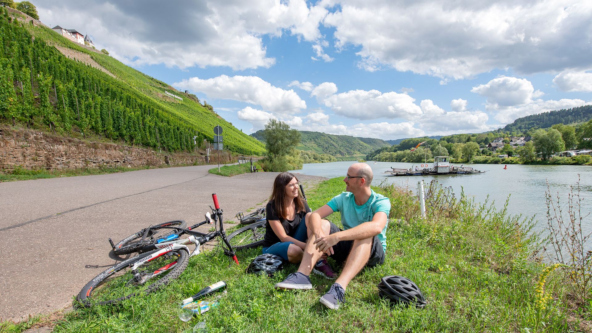 Radfahren Mosel Das Bild zeigt ein Paar, welches auf einem Stück Wiese an der Mosel ein Glas Wein genießt. Neben Ihnen liegen Fahrräder. Sie machen wohl eine kleine Pause vom Radfahren.