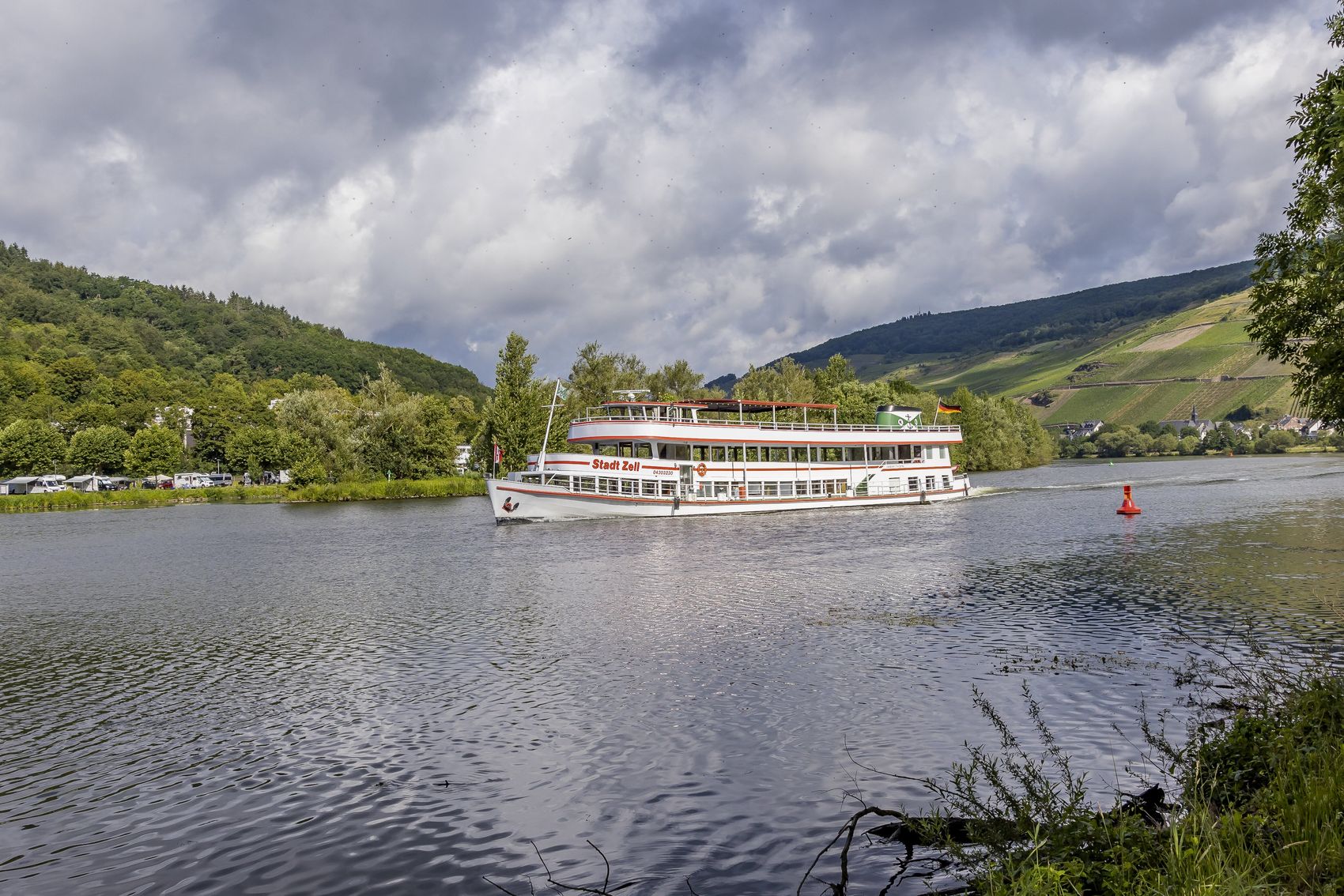 Schiff Das Bild zeigt das Schiff Stadt Zell auf der Mosel zwischen Zell-Merl und Zell-Kaimt bei bewölktem Wetter. Unten recchts in der Ecke befinden sich Sträucher vom Moselufer. Im Hintergrund erkennt man Weinberge und Wälder.
